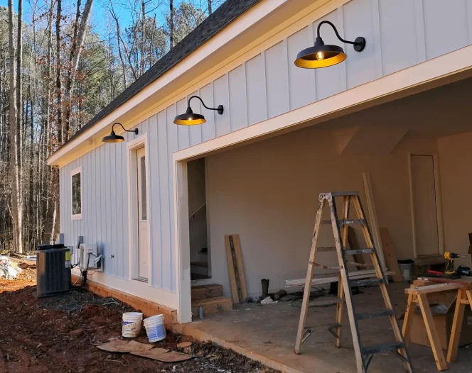 A newly built house with light gray vertical siding and three black barn lights with warm glowing bulbs.